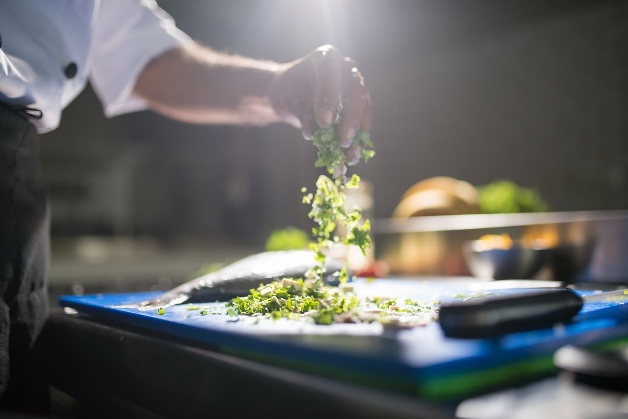 hero-img-01 Chef's hand preparing chopped green vegetables on a cutting board in a kitchen.