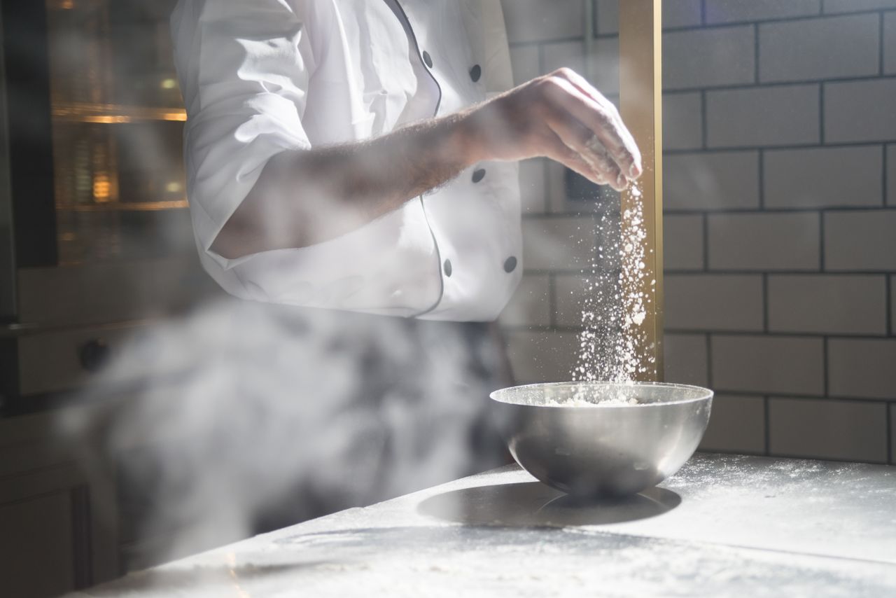 our-services-1 Chef in white uniform sprinkling flour into a steel bowl in a modern kitchen.